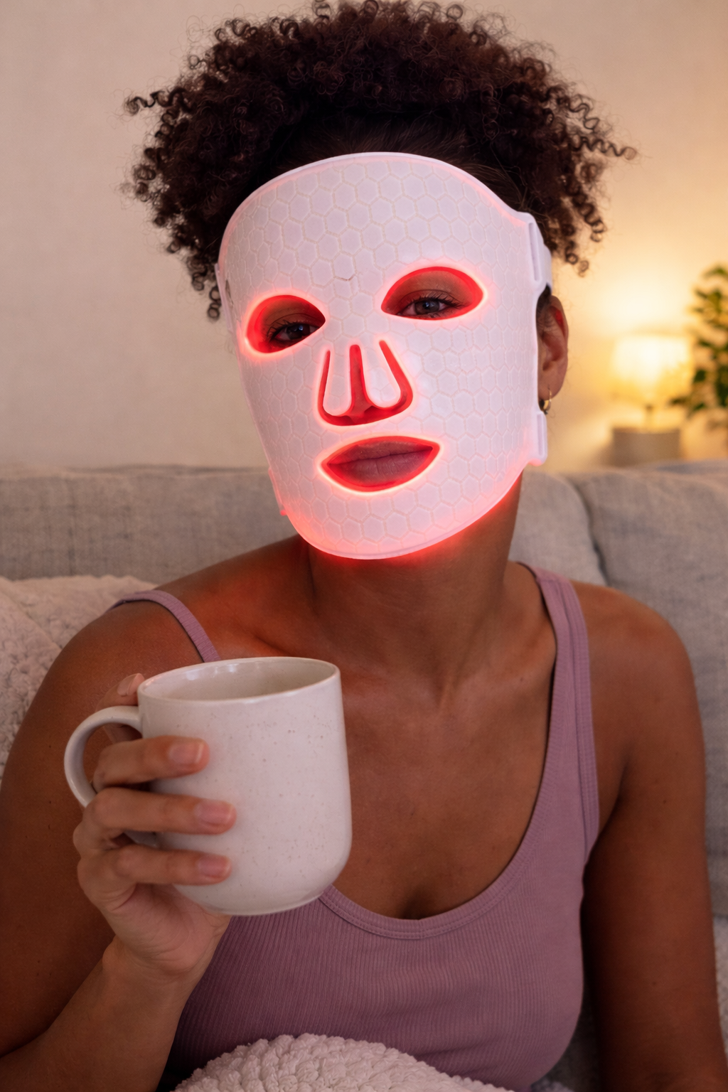 Person wearing a LED face mask holding a mug indoors.